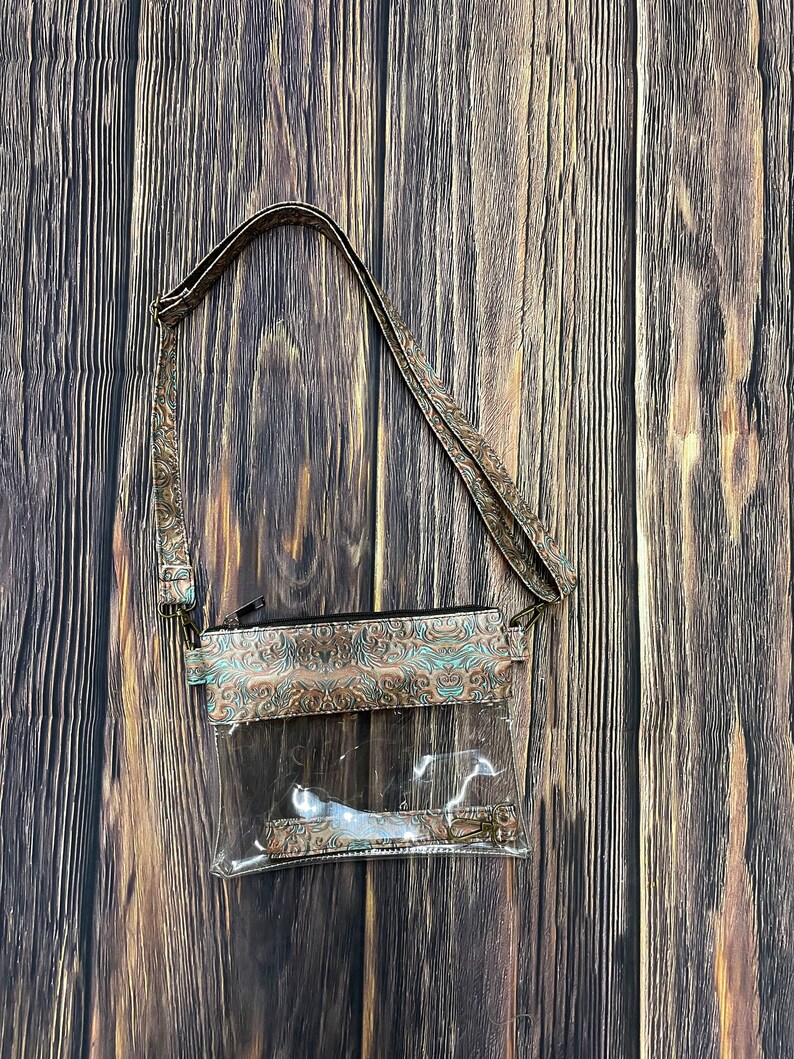 May include: A clear plastic crossbody bag with a brown and teal patterned fabric flap. The bag has a zipper closure and a long adjustable strap.