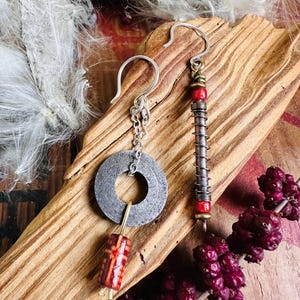 May include: Artisan earrings featuring a silver-tone metal washer with a chain and a red and brown bead. The other earring has a silver hook, red beads, and a metal coil design. The earrings are displayed on a piece of wood.