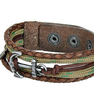 Puede incluir: Un brazalete de cuero marrón y cuerda con un dije de ancla de plata. El brazalete tiene tres capas de cuerda y una correa de cuero con dos broches de presión plateados.