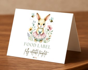 Puede incluir: Una tarjeta blanca plegada con una ilustración de acuarela de un conejito con tutú, rodeado de flores y abejas. El texto "FOOD LABEL" y "fully editable template" está impreso en la tarjeta. La tarjeta está sobre una superficie de madera.
