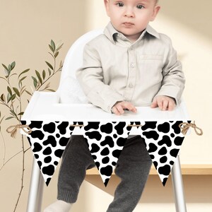 May include: A black and white cow print banner with triangular flags, strung together with twine. The banner is hanging in front of a baby sitting in a white high chair. The baby is wearing a light-coloured button-down shirt and grey trousers.
