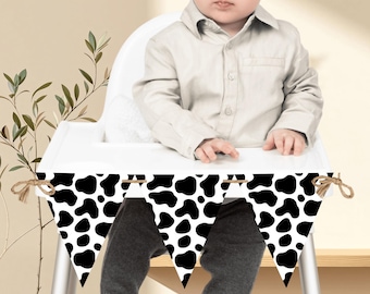 Peut inclure: Une guirlande de fanions triangulaires noirs et blancs à imprimé vache, attachés avec de la ficelle. La guirlande est suspendue devant un bébé assis dans une chaise haute blanche. Le bébé porte une chemise boutonnée de couleur claire et un pantalon gris.