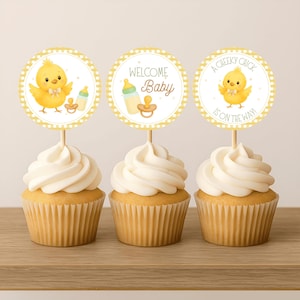 Puede incluir: Tres cupcakes con glaseado blanco y envoltorios amarillos. Cada cupcake tiene un adorno redondo con un diseño de pollito amarillo y el texto "Welcome Baby" y "A Cheeky Chick is on the Way!" Los adornos están sobre palitos de madera.