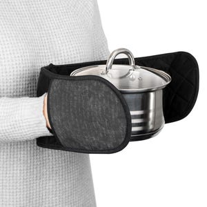 Puede incluir: Un agarrador negro con una superficie texturizada de color gris oscuro sostiene una olla de acero inoxidable con tapa de cristal. El agarrador tiene un diseño acolchado en la parte posterior. La olla la sostiene una persona que lleva un jersey gris.