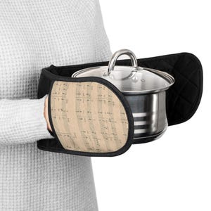 May include: Black oven mitts with a musical note design holding a stainless steel pot with a glass lid. The mitts have a quilted texture and a beige background with musical notes. The pot has a silver handle.