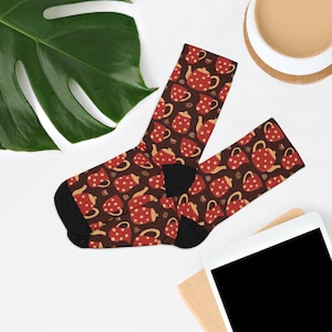 May include: A pair of socks with a brown background and a pattern of red teapots, mugs, and coffee beans with white polka dots. The socks have black heels and toes. The socks are on a white surface with a green leaf and a cup of coffee in the background.