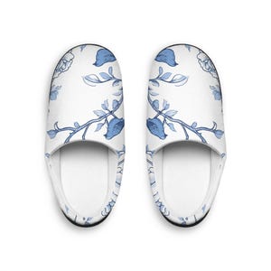 Peut inclure: Une paire de pantoufles blanches avec un motif floral bleu. Les pantoufles présentent un motif répétitif de fleurs et de feuilles bleues sur fond blanc. L'intérieur est blanc et les semelles sont noires.