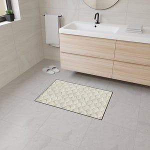 Puede incluir: Una alfombra de baño rectangular de color crema con un diseño tejido y un borde negro. La alfombra está colocada sobre un suelo de baldosas gris claro en un baño, cerca de un mueble de tocador de madera y un lavabo.