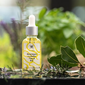 Puede incluir: Una botella de vidrio transparente con un gotero plateado y una etiqueta amarilla que dice "Rosemary Laurel True Oil". La botella está sobre una superficie negra con hojas verdes y flores de lavanda.