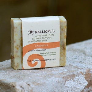 Puede incluir: Una pastilla de jabón natural con una etiqueta blanca que dice "KALLIOPE'S AGNO PURE LOCAL SAPOUNI OLIVE OIL ELAIOLADOU SOAP CALENDULA  μα πέταλα καλέντουλας handcrafted in Lefkas with local organic ingredients COLD PROCESS VEGAN PALM FREE".