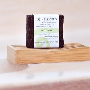 Może przedstawiać: Brązowe mydło z białą etykietą, na kt&oacute;rej widnieje napis "KALLIOPE'S AGNO PURE LOCAL SAPOUNI OLIVE OIL ELAIOLADOU SOAP ECO COFFEE &alpha;&pi;&omicron;&lambda;&epsilon;&pi;&iota;&sigma;&tau;&iota;&kappa;ό &mu;&epsilon; &kappa;&alpha;&phi;έ handcrafted in Lefkos with 100% organic ingredients COLD PROCESS VEGAN PALM FREE". Mydło leży na drewnianej mydelniczce.