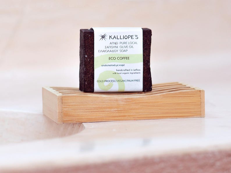 Może przedstawiać: Brązowe mydło z białą etykietą, na kt&oacute;rej widnieje napis "KALLIOPE'S AGNO PURE LOCAL SAPOUNI OLIVE OIL ELAIOLADOU SOAP ECO COFFEE &alpha;&pi;&omicron;&lambda;&epsilon;&pi;&iota;&sigma;&tau;&iota;&kappa;ό &mu;&epsilon; &kappa;&alpha;&phi;έ handcrafted in Lefkos with 100% organic ingredients COLD PROCESS VEGAN PALM FREE". Mydło leży na drewnianej mydelniczce.