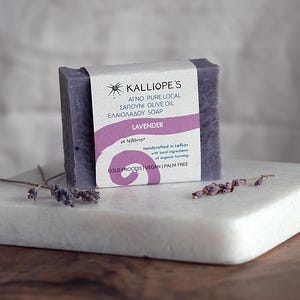 Puede incluir: Una pastilla de jabón rectangular de color lavanda con una etiqueta blanca y detalles morados, sobre una bandeja de mármol blanco. La etiqueta dice "Kalliope's Lavender Soap". Ramitas de lavanda seca están cerca del jabón.