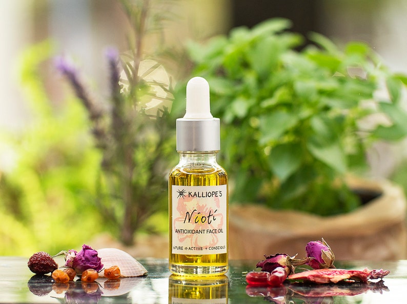May include: A glass bottle of Nioti Antioxidant Face Oil by Kalliope's. The bottle is clear and has a gold label with the product name and description. The bottle is sitting on a table with a few dried flowers and a seashell.