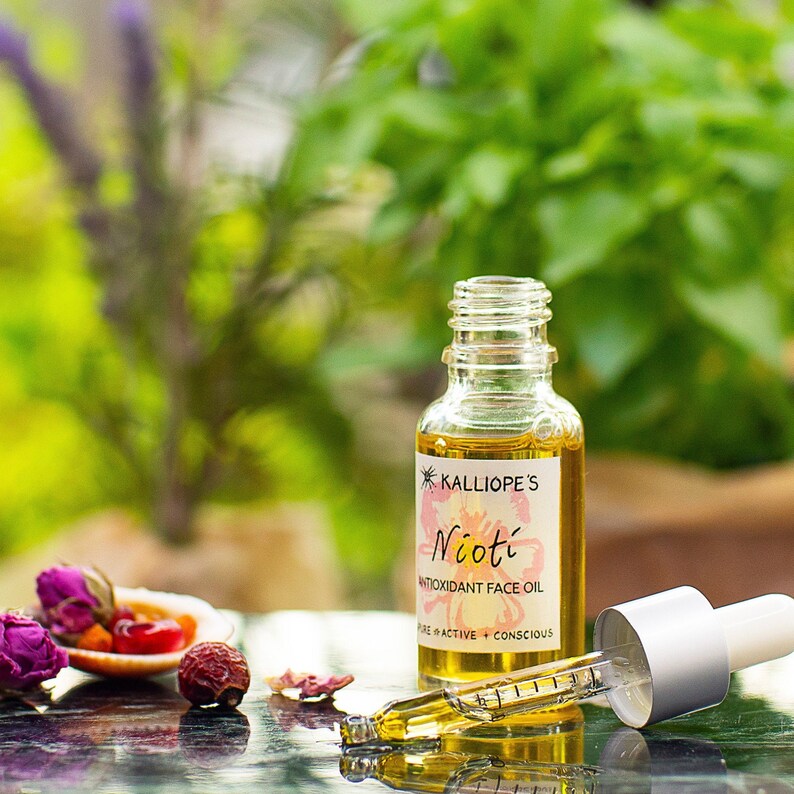 May include: A glass bottle of Nioti Antioxidant Face Oil by Kalliope's. The bottle is clear and has a textured surface. The label is white with black text and a pink flower illustration. The bottle is sitting on a table with a dropper next to it.