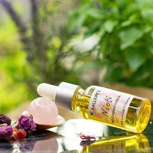 May include: A small glass bottle of face oil labeled "Kalliope's Nioti Antioxidant Face Oil" with a pink rose and a rose quartz crystal on a black surface.