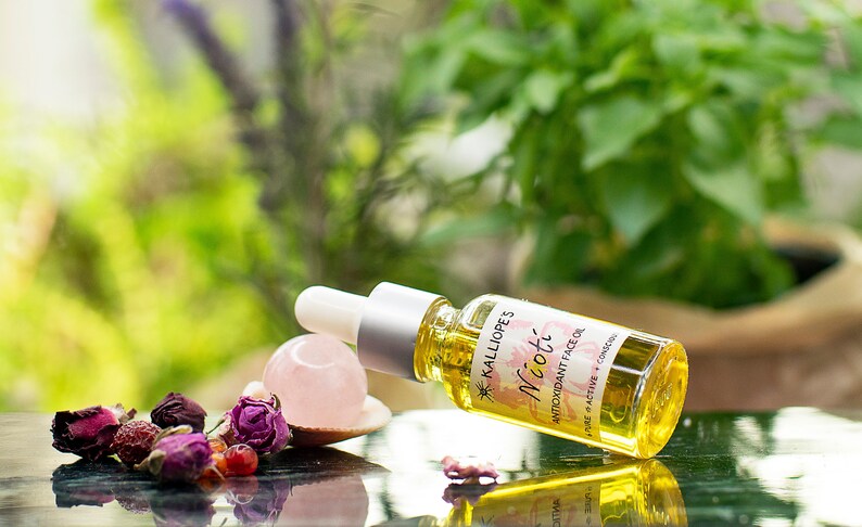 May include: A small glass bottle of face oil labeled "Kalliope's Nioti Antioxidant Face Oil" with a pink rose and a rose quartz crystal on a black surface.