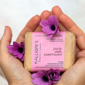 May include: A brown paper-wrapped bar of solid hair conditioner with the brand name "KALLIOPE'S" and the text "SOLID HAIR CONDITIONER" printed on the front. The bar is surrounded by purple flowers.