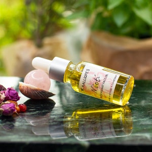 May include: A clear glass bottle with a dropper top, labeled "KALLIOPE'S Nicot ANTIOXIDANT FACE OIL PRE-ACTIVE + CONSCIOUS". The bottle is lying on a green surface with a pink crystal and a seashell.