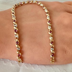 May include: A gold-tone tennis bracelet featuring a repeating twisted metal design. The bracelet is set with alternating round-cut red rubies and clear white diamonds or cubic zirconia stones arranged in a continuous line with a secure metal clasp.
