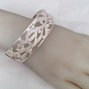 May include: A silver bangle bracelet with a delicate, intricate design featuring a floral pattern. The bracelet is adorned with clear cubic zirconia stones.
