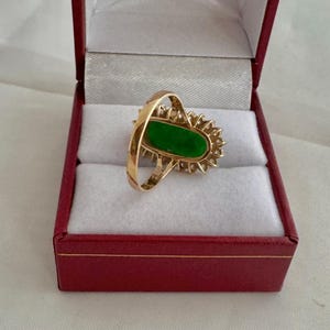 May include: A gold ring with a green oval gemstone surrounded by a halo of smaller diamonds. The ring is displayed in a red velvet ring box.