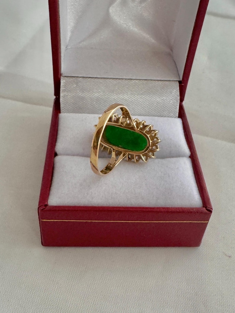 May include: A gold ring with a green oval gemstone surrounded by a halo of smaller diamonds. The ring is displayed in a red velvet ring box.