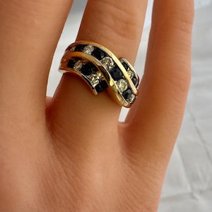 May include: A gold ring with a wave design, set with alternating rows of dark blue and clear gemstones. The ring is on a finger, showcasing the intricate details of the jewelry.
