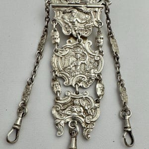May include: A silver pocket watch chain with intricate designs and three chains. The chain features a series of ornate, interlocking links and decorative elements.
