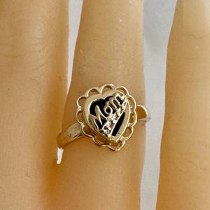 May include: A gold-toned ring featuring a heart-shaped design with the word "Mom" in a stylized font. The heart is framed by a scalloped border. The ring is on a finger.