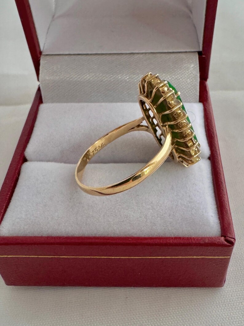 May include: A gold ring with a large green gemstone surrounded by smaller diamonds. The ring is in a red box with white lining.