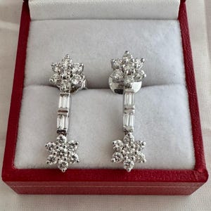 May include: A pair of diamond earrings displayed in a red jewelry box. Each earring features a cluster of round diamonds at the top and bottom, connected by a vertical bar of baguette-cut diamonds. The earrings are set in white metal.
