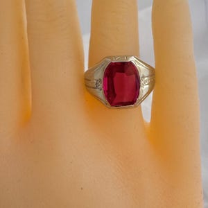 May include: A gold ring with a large, red, rectangular gemstone set in a square bezel. The ring has a detailed, engraved design.