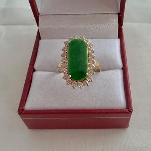 May include: A gold ring with a large green jade stone surrounded by a halo of small clear stones. The ring is in a red velvet ring box.