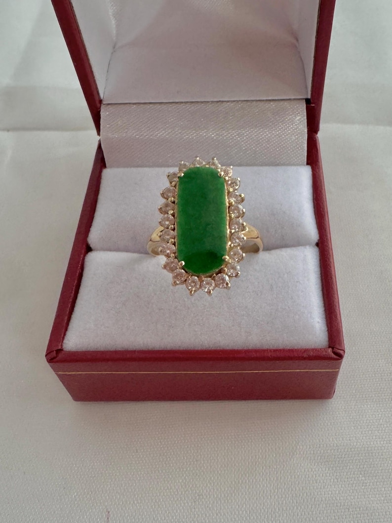 May include: A gold ring with a large green jade stone surrounded by a halo of small clear stones. The ring is in a red velvet ring box.