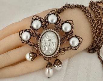 May include: An antique-style necklace with a bronze-colored chain and pendant. The pendant features a central oval cameo surrounded by faux pearls and a filigree design. Additional faux pearl drops hang from the bottom.