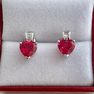 May include: A pair of heart-shaped ruby earrings set in silver. The earrings are displayed in a red jewelry box with a white interior. The rubies are a vibrant red color and are cut into a heart shape.