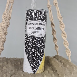 May include: A black and white composition book themed tumbler with the text "Composition Book" and "Mrs. Alford" on the front. The tumbler is hanging from a macrame rope.