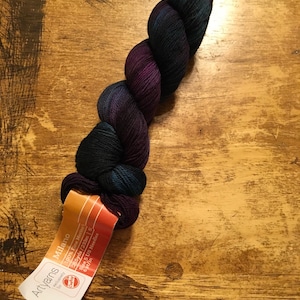 May include: A skein of Milano 100% Fine Merino yarn in shades of dark teal, purple, and black. The yarn is twisted and has a label attached with the Artyarns logo and product details. The label indicates the yarn is 233 meters and recommends size 5 US needles.