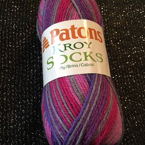 Può includere: Un gomitolo di filato Patons Kroy Socks nei toni del viola, rosa e grigio. Il filato è avvolto con un'etichetta bianca che recita "Patons KROY SOCKS". Il filato è progettato per la lavorazione a maglia di calzini.