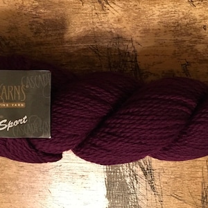 Op de afbeelding: Een streng Cascade 220 Sport garen in een diepe bordeauxrode kleur. Het garen is strak opgerold en heeft een zachte, getextureerde uitstraling. Het etiket leest "Cascade Yarns" en "Sport".
