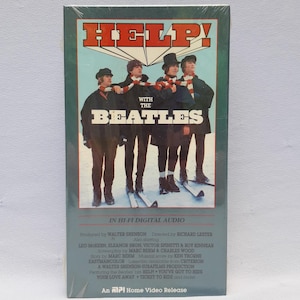 May include: A vintage VHS tape of "Help!" with The Beatles. The cover features the band in a group pose with the title in bold letters. The tape is in its original packaging, with text at the bottom indicating it's a home video release.