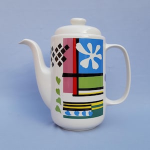 Pode incluir: Bule de cerâmica branco com tampa e alça, apresentando um design geométrico colorido. O design inclui diamantes pretos, retângulos rosa e vermelhos, uma flor azul, listras verdes e amarelas e corações verdes. O bule está sobre um fundo azul claro.