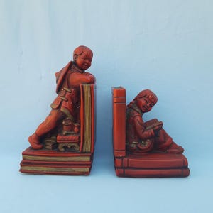Puede incluir: Par de sujetalibros rojos que representan a niños con vestimenta tradicional. Uno se apoya en una pila de libros, el otro lee. Los detalles dorados resaltan las figuras y los libros. Un complemento decorativo para cualquier estantería.