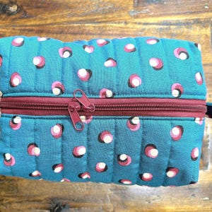 May include: A teal quilted pouch with a repeating pattern of pink, white, and black polka dots. The pouch has a burgundy zipper and a small fabric loop on the side. The design is symmetrical and the stitching is visible.