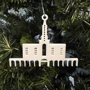 Puede incluir: Adorno blanco de plástico con forma de iglesia con una aguja y una parte superior redondeada. El adorno tiene una serie de arcos en la base.