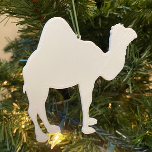 May include: White ceramic camel ornament hanging on a green Christmas tree. The ornament is a simple silhouette of a camel.