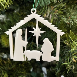 May include: White plastic Christmas ornament depicting the nativity scene. The ornament features a manger with a star above it, a figure of Mary kneeling beside the manger, and a figure of Joseph standing beside Mary.