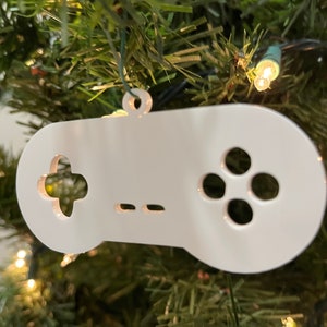 Puede incluir: Adorno navideño de plástico blanco con forma de mando de videojuego. El adorno tiene un botón en forma de cruz, dos botones más pequeños y una almohadilla circular de botones.