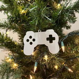 Puede incluir: Adorno navideño de plástico blanco con forma de mando de videojuego. El adorno está colgado de un árbol de Navidad verde con luces blancas.